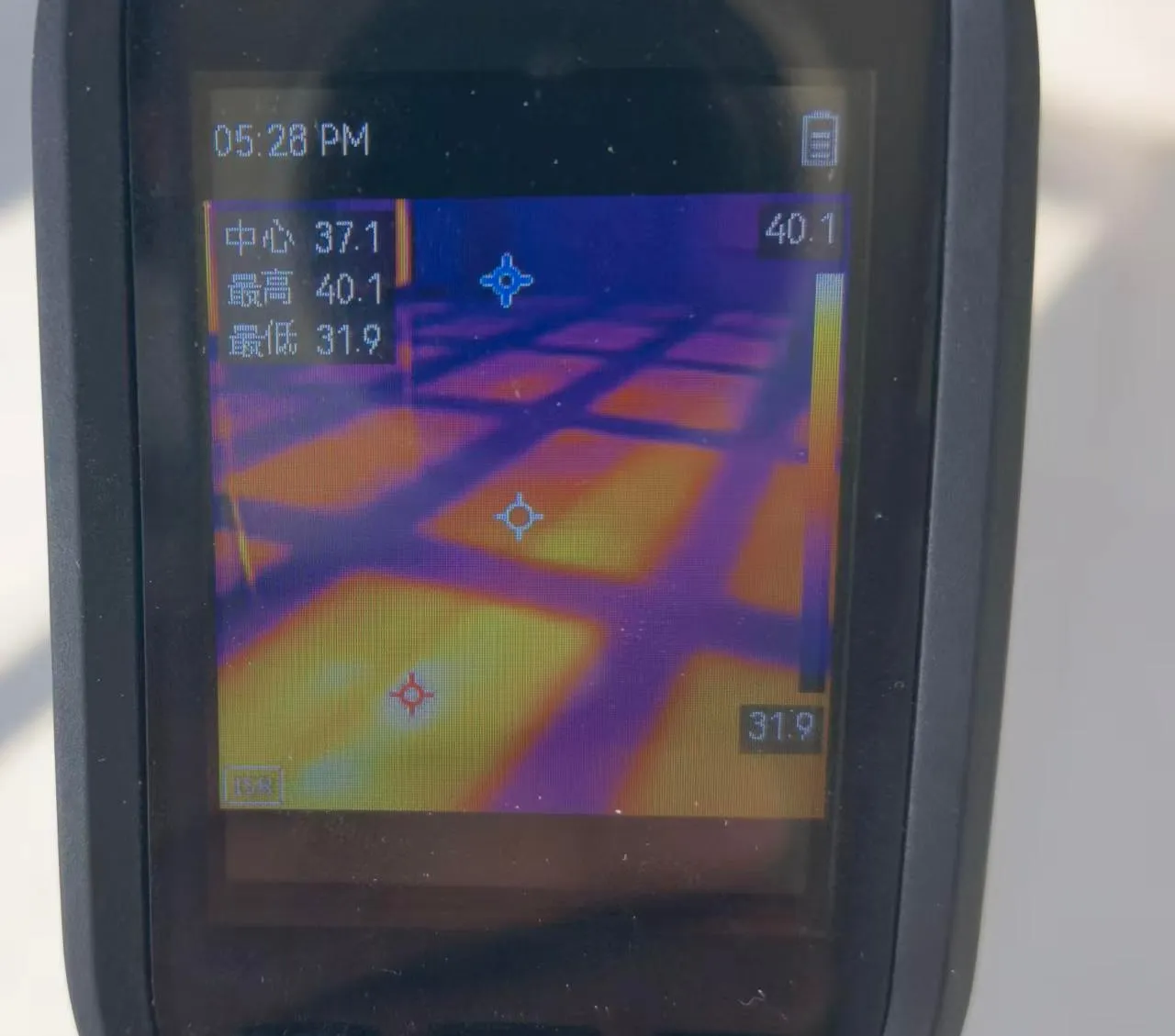 A thermal camera held over a heated floor, its screen showing each heater glowing evenly at 37 °C